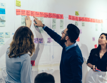 Female with dark hair writing against a poster pinned to a board during a consultancy workshop delivered by IfM Engage.